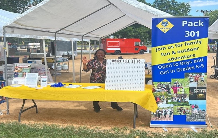 An adult volunteer waving from behind a booth with materials laid out on the table